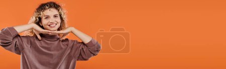fröhliche Frau im braunen Rollkragen mit den Händen unterm Kinn, die in die Kamera auf orangefarbenem Banner blickt