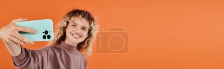 sonriente mujer de moda con el pelo ondulado tomar selfie en el teléfono inteligente en fondo naranja borrosa, bandera