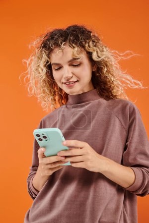 pleased curly woman in brown turtleneck messaging on mobile phone on orange studio backdrop