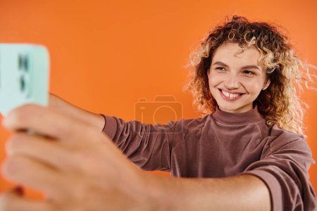 mujer excitada con el pelo ondulado tomando selfie en el teléfono móvil borroso en el fondo naranja radiante