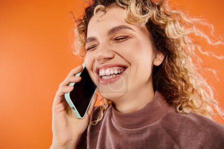 überglücklich lockige Frau lacht während Handy-Gespräch auf leuchtend orangefarbenem Hintergrund, Glück