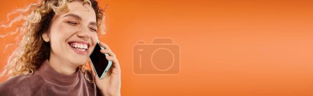 Photo for Cheerful woman with wavy hair laughing during conversation on smartphone on orange backdrop, banner - Royalty Free Image