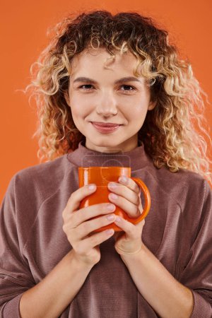 Téléchargez les photos : Femme souriante avec les cheveux bouclés et tasse de café du matin en regardant la caméra sur orange, routine quotidienne - en image libre de droit
