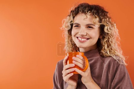 fröhliche Frau mit lockigem Haar und einer Tasse Morgenkaffee, die auf orangefarbene, tägliche Routine blickt
