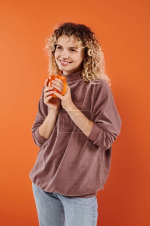 joyful woman with curly hair and cup of morning coffee looking away on orange, daily routine