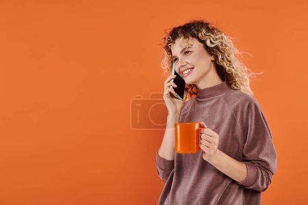 cheerful curly woman with cup of morning coffee talking on smartphone on bright orange backdrop