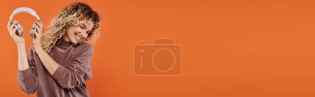 Téléchargez les photos : Femme joyeuse et à la mode en couleur moka col roulé tenant écouteurs sans fil sur orange, bannière - en image libre de droit