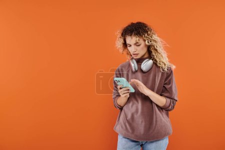 Foto de Mujer rizada con estilo con la mensajería de auriculares inalámbricos en el teléfono móvil en fondo naranja - Imagen libre de derechos