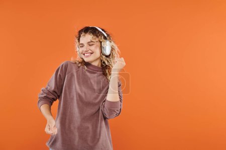 Téléchargez les photos : Femme excitée avec des cheveux ondulés dansant dans des écouteurs sans fil et couleur moka col roulé sur orange - en image libre de droit