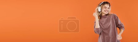 Téléchargez les photos : Femme joyeuse avec des cheveux ondulés dansant dans les écouteurs et le col roulé couleur moka sur orange, bannière - en image libre de droit