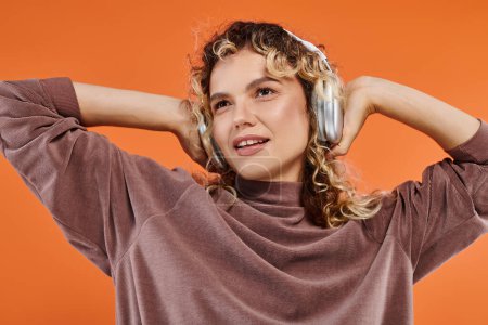 impressed curly in brown turtleneck woman listening music in wireless headphones on orange backdrop