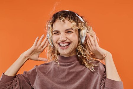 mujer rizada de moda y muy alegre en auriculares inalámbricos escuchando música sobre fondo naranja