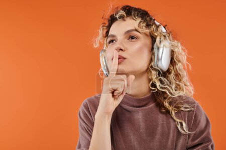 Téléchargez les photos : Femme attrayante avec des cheveux ondulés écouter de la musique dans les écouteurs sans fil montrant signe de silence sur orange - en image libre de droit