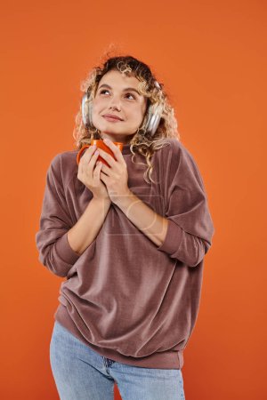 Foto de Mujer rizada de ensueño con taza de café de la mañana escuchando música en auriculares inalámbricos en naranja - Imagen libre de derechos