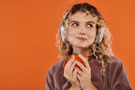 verträumte, glückliche Frau mit einer Tasse Morgenkaffee und Musik in drahtlosen Kopfhörern auf orange