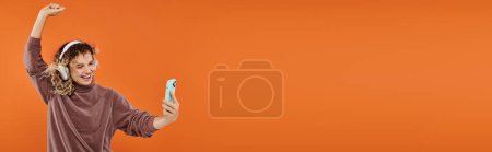 Photo for Excited curly woman dancing in wireless headphones and taking selfie on smartphone on orange, banner - Royalty Free Image