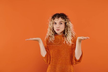 confused curly woman in knitted sweater puffing cheeks and showing shrug gesture on orange