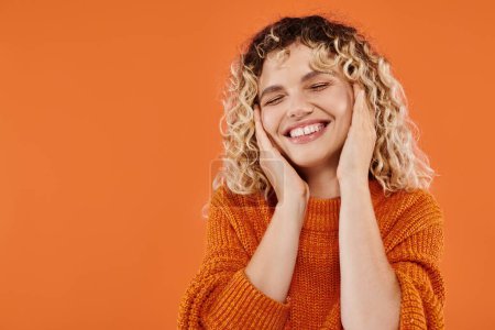 Lockige aufgeregte Frau im hellen Pullover lacht mit geschlossenen Augen vor leuchtend orangefarbenem Hintergrund