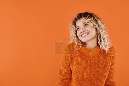mujer feliz y rizada en suéter de punto brillante sonriendo mirando hacia otro lado en el fondo del estudio naranja