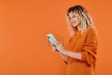 Lächeln lockige Frau in hellem Strickpullover surft im Internet auf Smartphone auf orangefarbenem Hintergrund