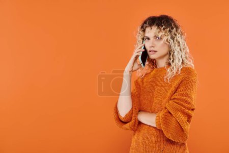 ernste lockige Frau in stylischem Strickpullover, die auf dem Handy vor leuchtend orangefarbenem Hintergrund spricht