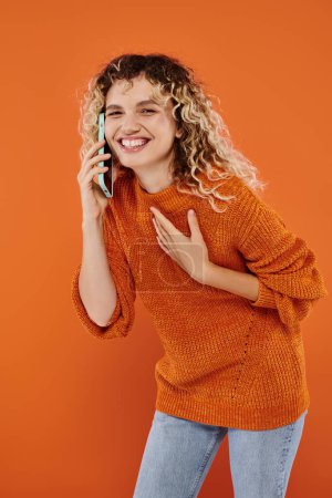 überglückliche lockige Frau im Strickpullover, die mit dem Handy telefoniert und auf orangefarbenem Hintergrund lacht