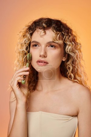 Téléchargez les photos : Charmante femme aux cheveux bouclés et à la peau parfaite en utilisant un rouleau de jade sur fond rose et jaune - en image libre de droit