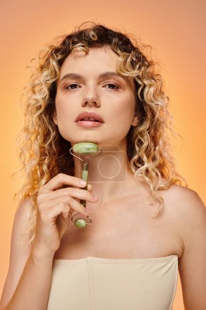sensual curly woman with radiant skin using jade roller and looking at camera on pastel backdrop