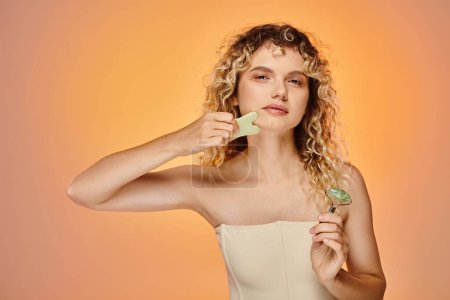 charming curly woman with jade roller and gua sha looking at camera on pink and yellow backdrop