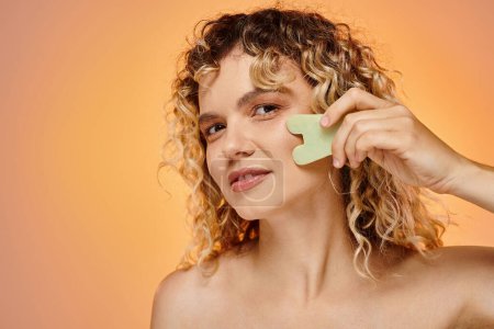happy curly woman with perfect skin using gua sha looking at camera on pastel gradient backdrop