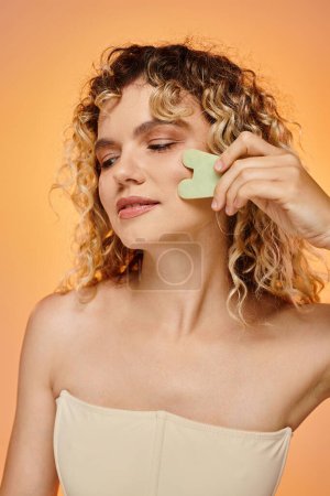 sensual woman with wavy hair and glowing skin using gua sha on pink and yellow gradient backdrop