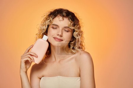 pleased woman with wavy hair and glowing skin holding bottle of body lotion on pastel backdrop