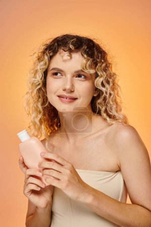 fröhliche Frau mit lockigem Haar hält Flasche mit Körperlotion auf pastellfarbenem Hintergrund