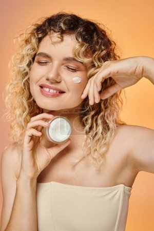 erfreut Frau mit lockigem Haar und sauberer Haut hält Glas Gesichtscreme auf gelb und rosa Hintergrund