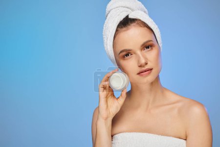 alluring woman with towel on head showing jar of face cream on blue backdrop, wellness and beauty Poster 696260042