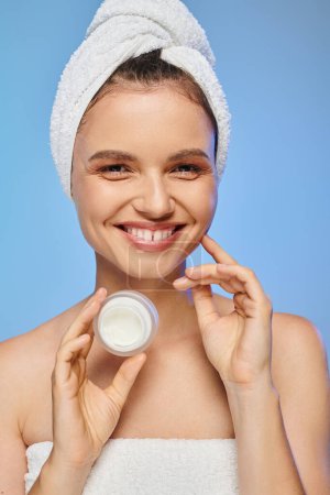 cheerful woman with towel on head holding jar of face cream on blue backdrop, wellness and beauty mug #696260174
