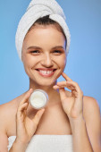 cheerful woman with towel on head holding jar of face cream on blue backdrop, wellness and beauty mug #696260174