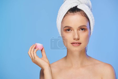 attractive woman with glowing skin and towel holding cosmetic cream and looking at camera on blue puzzle 696260410