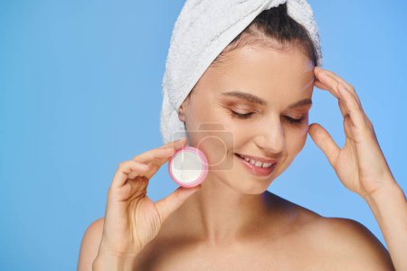 pleased charming woman with closed eyes and towel holding face cream and smiling on blue backdrop