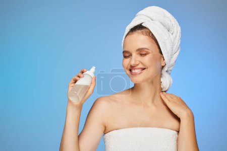 Photo for Cheerful woman with towel on head and perfect face holding dispenser with cosmetic foam on blue - Royalty Free Image