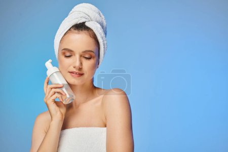 lächelnde Frau mit glühender Haut und Handtuch auf dem Kopf, Spender mit Gesichtsschaum auf blau