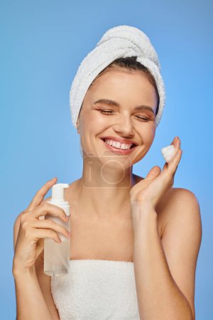 delighted woman with closed eyes and towel on head holding bottle of face foam on blue backdrop