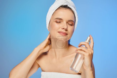 smiling woman with radiant skin and closed eyes holding bottle of body spray on blue backdrop