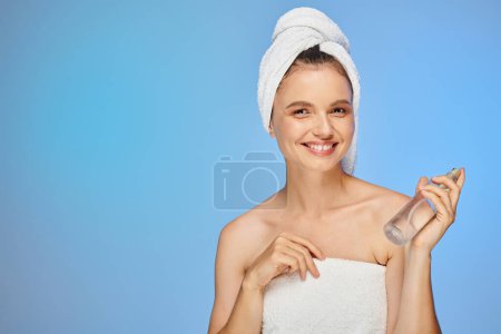 cheerful woman with towel on head and bottle of body spray looking at camera on blue backdrop