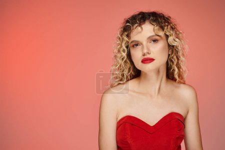 Photo for Fashionable curly woman with bold makeup in red top looking at camera on pink and yellow backdrop - Royalty Free Image
