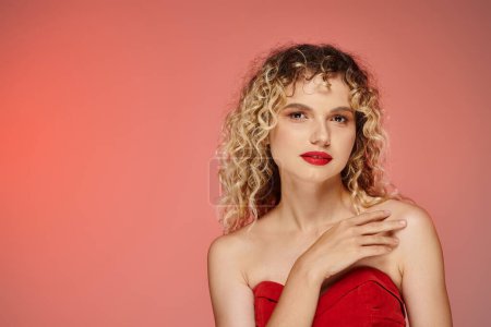 Téléchargez les photos : Femme bouclée sensuelle avec un maquillage audacieux en haut rouge regardant la caméra sur fond rose et jaune - en image libre de droit