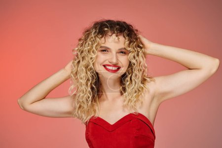 joyful woman with bold makeup in red top touching wavy hair and looking at camera on pastel backdrop