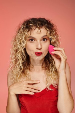 Téléchargez les photos : Femme étonnante avec les cheveux bouclés et les lèvres rouges en utilisant une éponge visage sur fond de dégradé rose et jaune - en image libre de droit