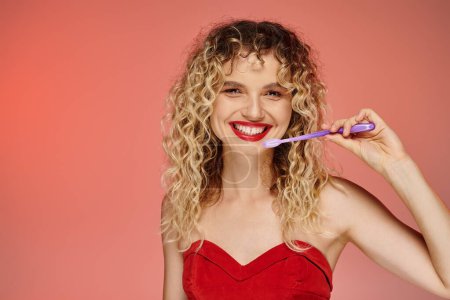 cheerful curly woman with red lips and toothbrush looking at camera on pastel gradient backdrop