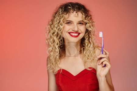 Foto de Mujer fascinante con el pelo ondulado y labios rojos sosteniendo cepillo de dientes y mirando a la cámara en rosa - Imagen libre de derechos